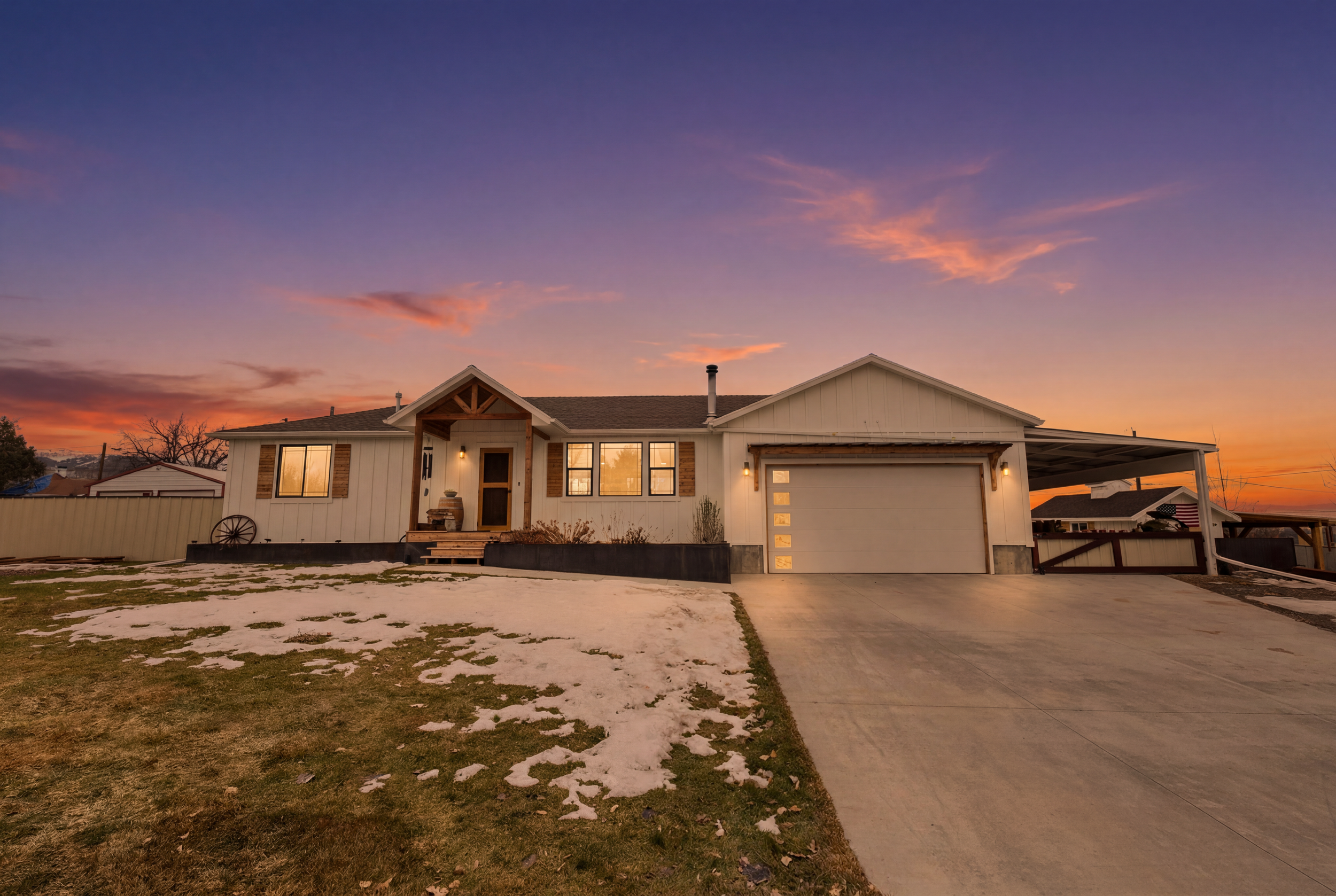 A suburban house at sunset with a white exterior, a porch, a closed garage door, and a partly frozen lawn.