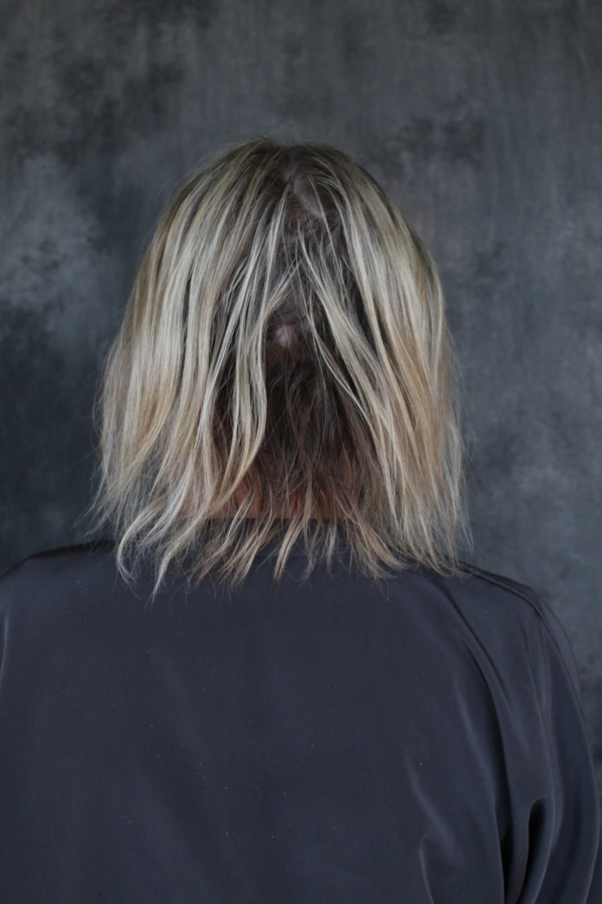 Back of a person with tousled, blonde hair and wearing a dark jacket, standing against a dark, textured background.