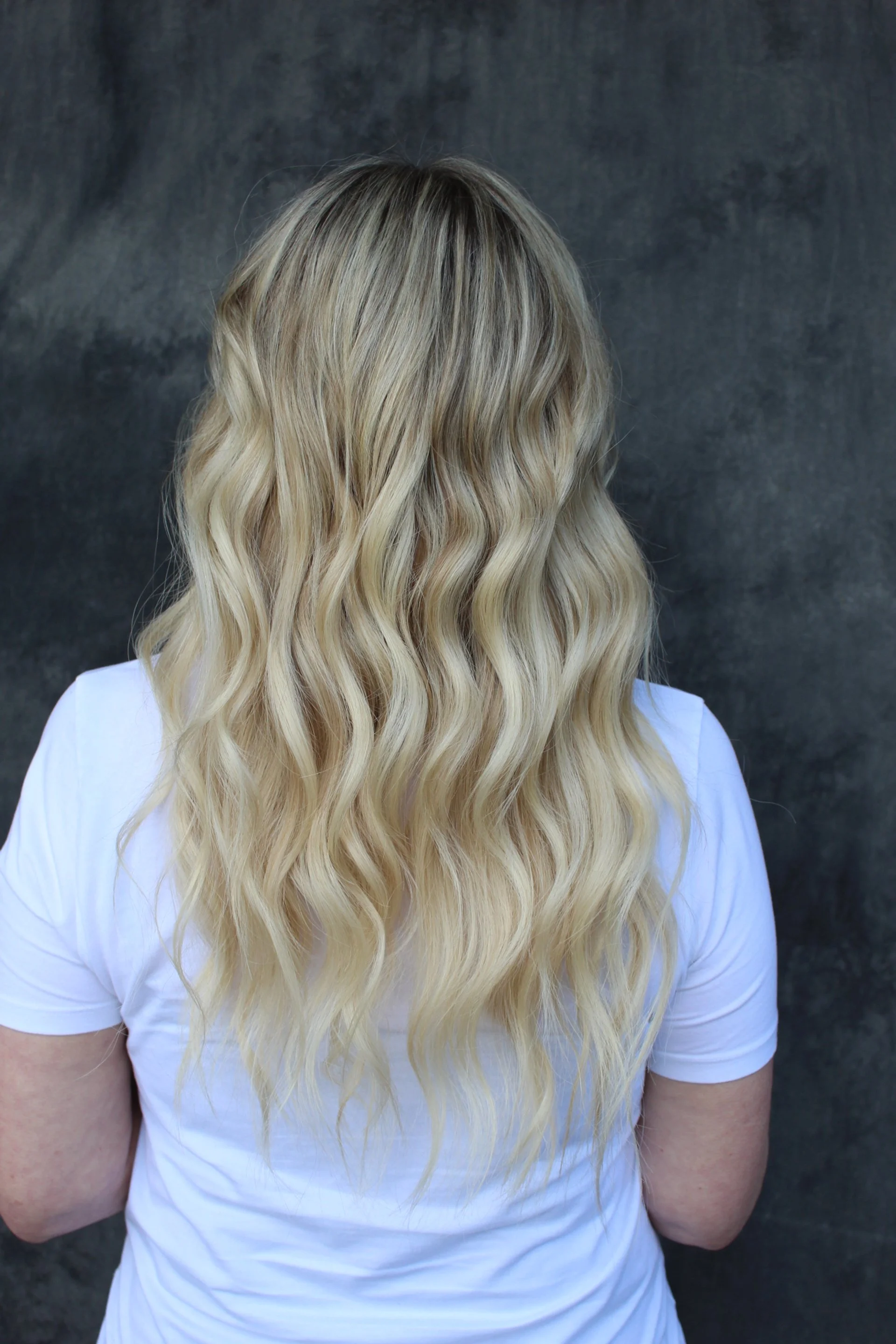 Back view of a woman with long, wavy blonde hair wearing a white t-shirt against a dark textured background.