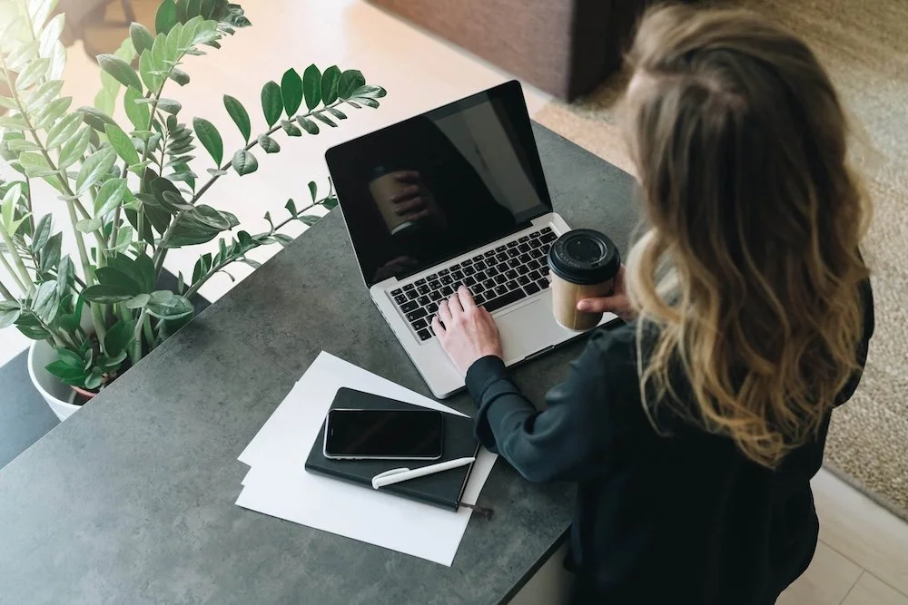A woman working on a laptop at a desk with a potted plant, smartphone, notebook, and coffee cup.