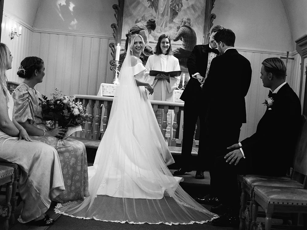Et svart-hvitt bilde av et bryllupsseremoni med bruden i hvit kjole og brudepassere, samt forlovere og brudepiker, i en pyntet rom med et religiøst maleri i bakgrunnen.