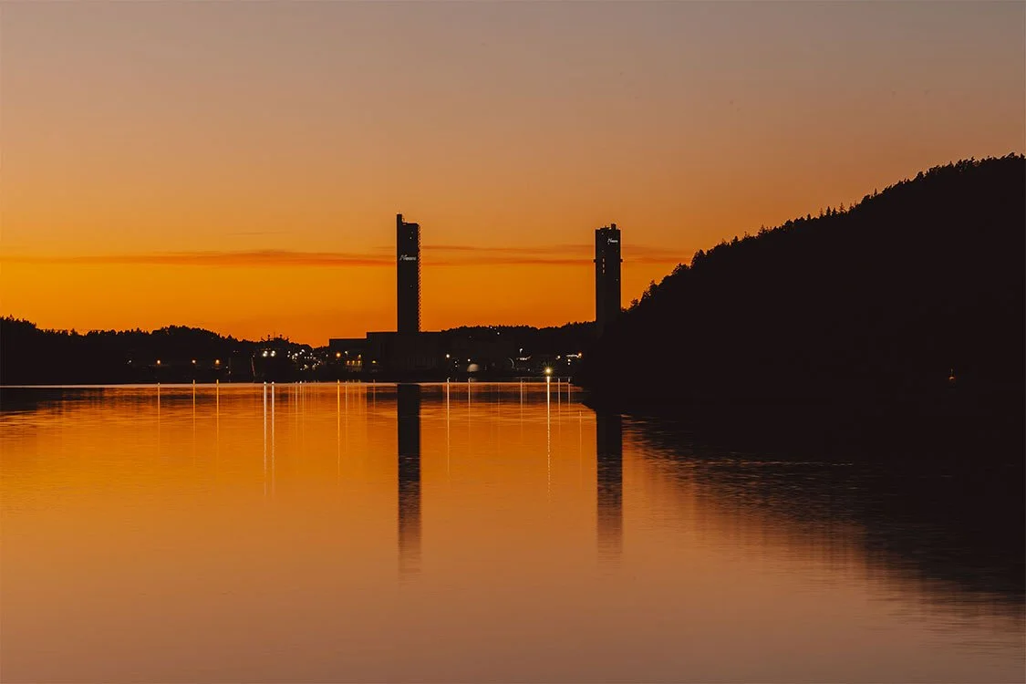 En fredelig innsjø ved solnedgang med to høye skyskrapere i bakgrunnen, og fjell på høyre side, reflektert i vannet.