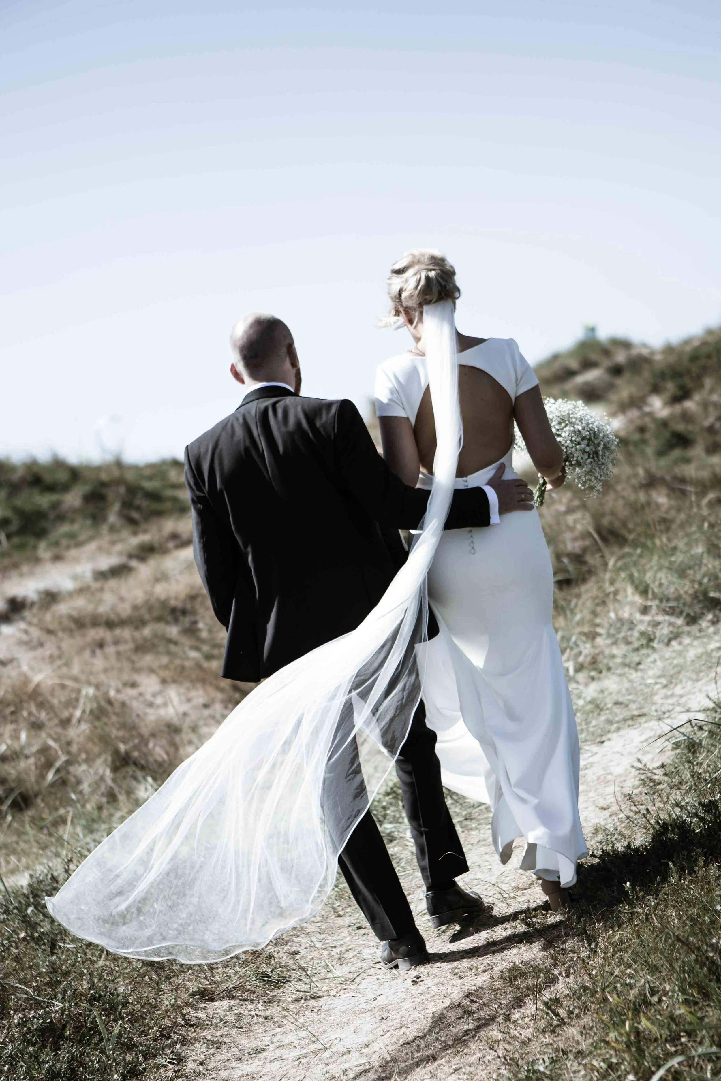 Brudepar går hånd i hånd på en sandvei i et naturskjønt område under klar himmel.