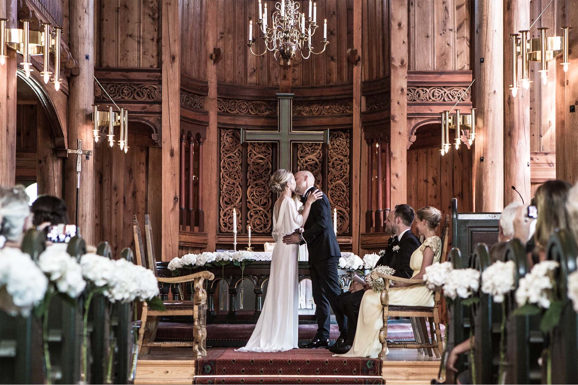 Et bryllup i en kirke med tre par, der alle kler seg i formelle antrekk, midt i seremoni foran en stor trekross. Brudeparet kysser, mens to andre par venter. Kledde i elegante farger, omgitt av blomster og treverk.