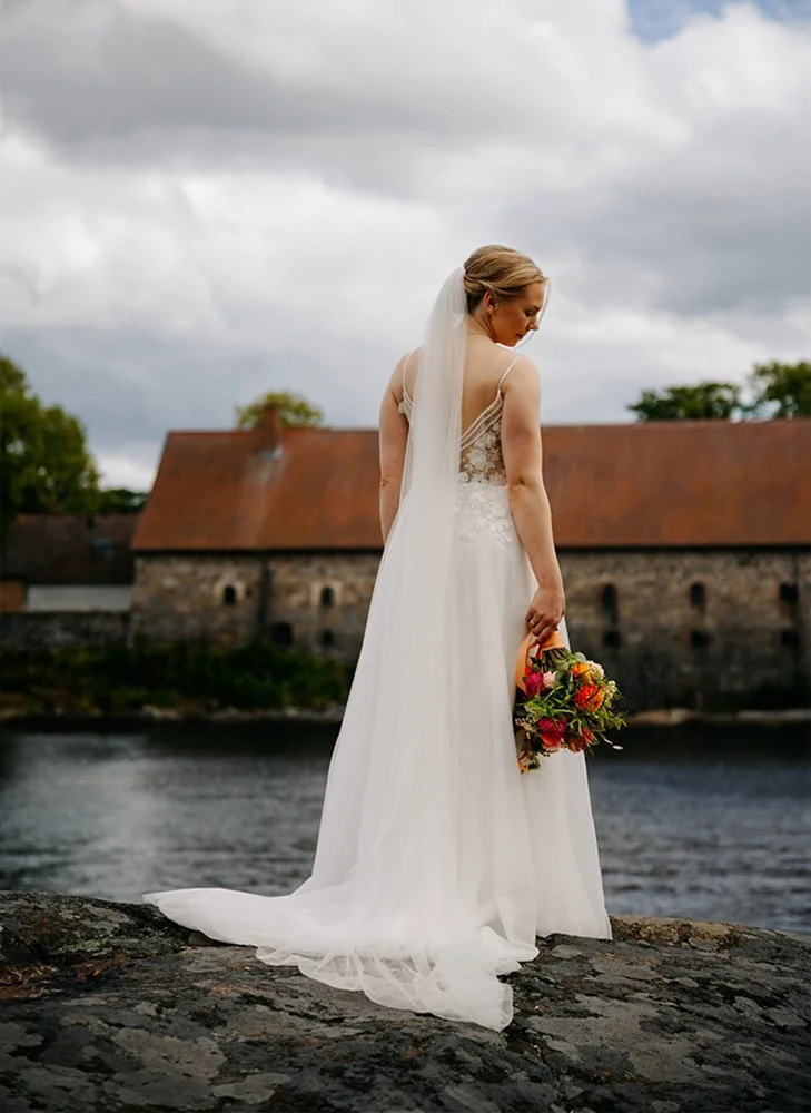 En brud i hvit brudekjole holder en blomsterbukett mens hun står på en stein nær en elv, med en gammel bygning med tak i bakgrunnen og overskyet himmel.