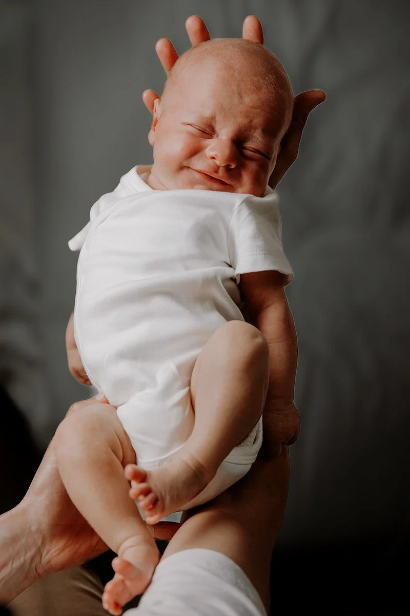 Nydelig nyfødt baby som holdes opp av en person, smiler med lukkede øyne, iført hvit t-skjorte.