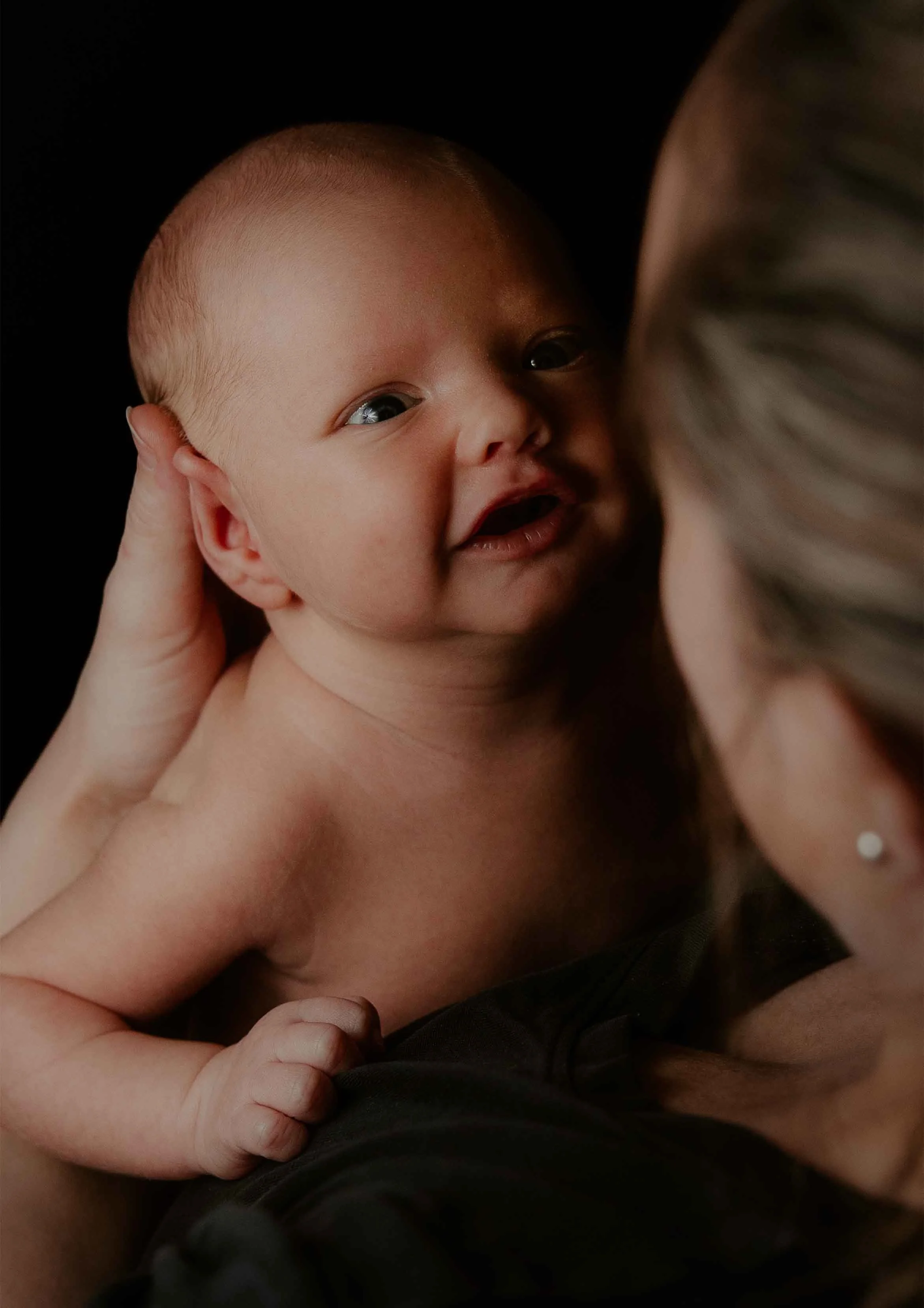 Nærbilde av en smilende baby som blir holdt av en voksen, mot en svart bakgrunn.