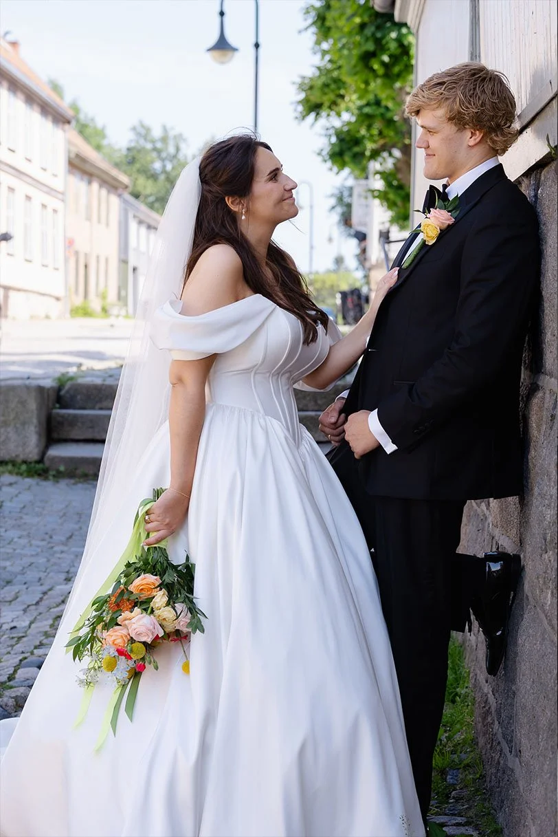 Et brudepar står inntil en mur på en gate. Bruden bærer en hvit kjole og holder en blomsterbukett, mens brudgommen har smoking med blomster i knapphullet.