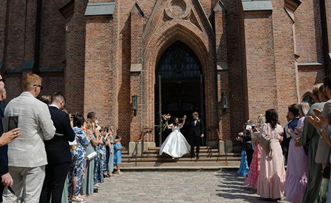 En brud og en brudgom utenfor en gammel, mursteinskirke, omringet av bryllupsgjester som applauderer.