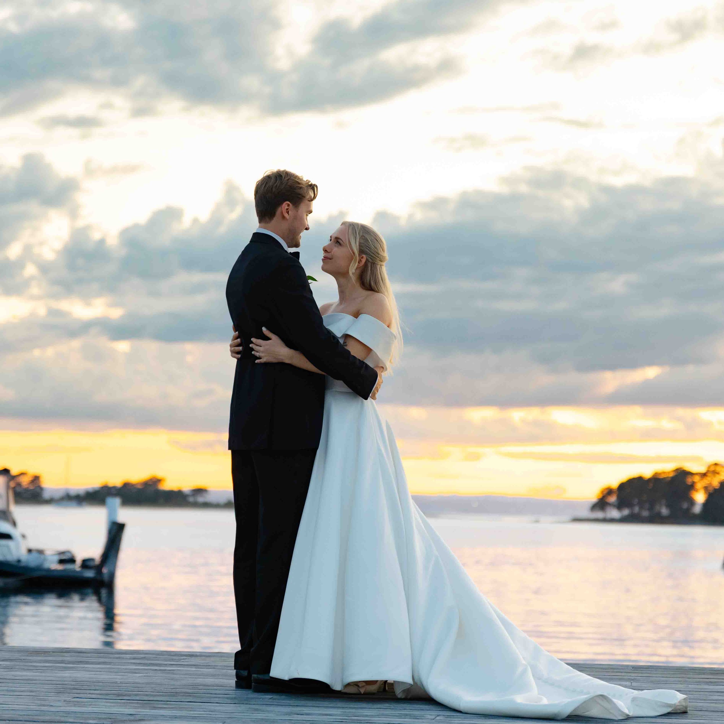 Et brudepar står ansikt til ansikt med havet i bakgrunnen under en solnedgang, og ser på hverandre med kjærlighet. Bruden har på seg en hvit brudekjole, og brudgommen har på seg en svart smoking.