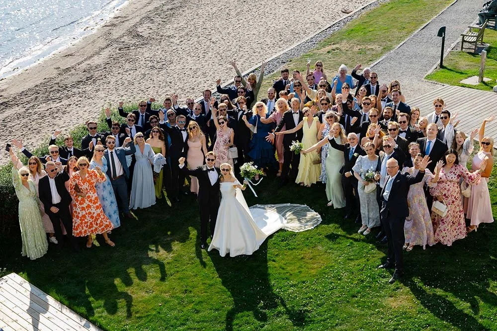 Et stort gruppebilde av brudeparet og gjester på en strand ved et bryllup, tatt fra luften.