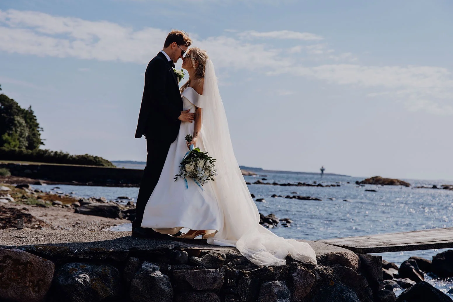 Et nygift par står på en stein på stranden med havet og en himmel med lette skyer i bakgrunnen. De ser på hverandre og holder hender, med bryllupsantrekk og blomsterbukett.