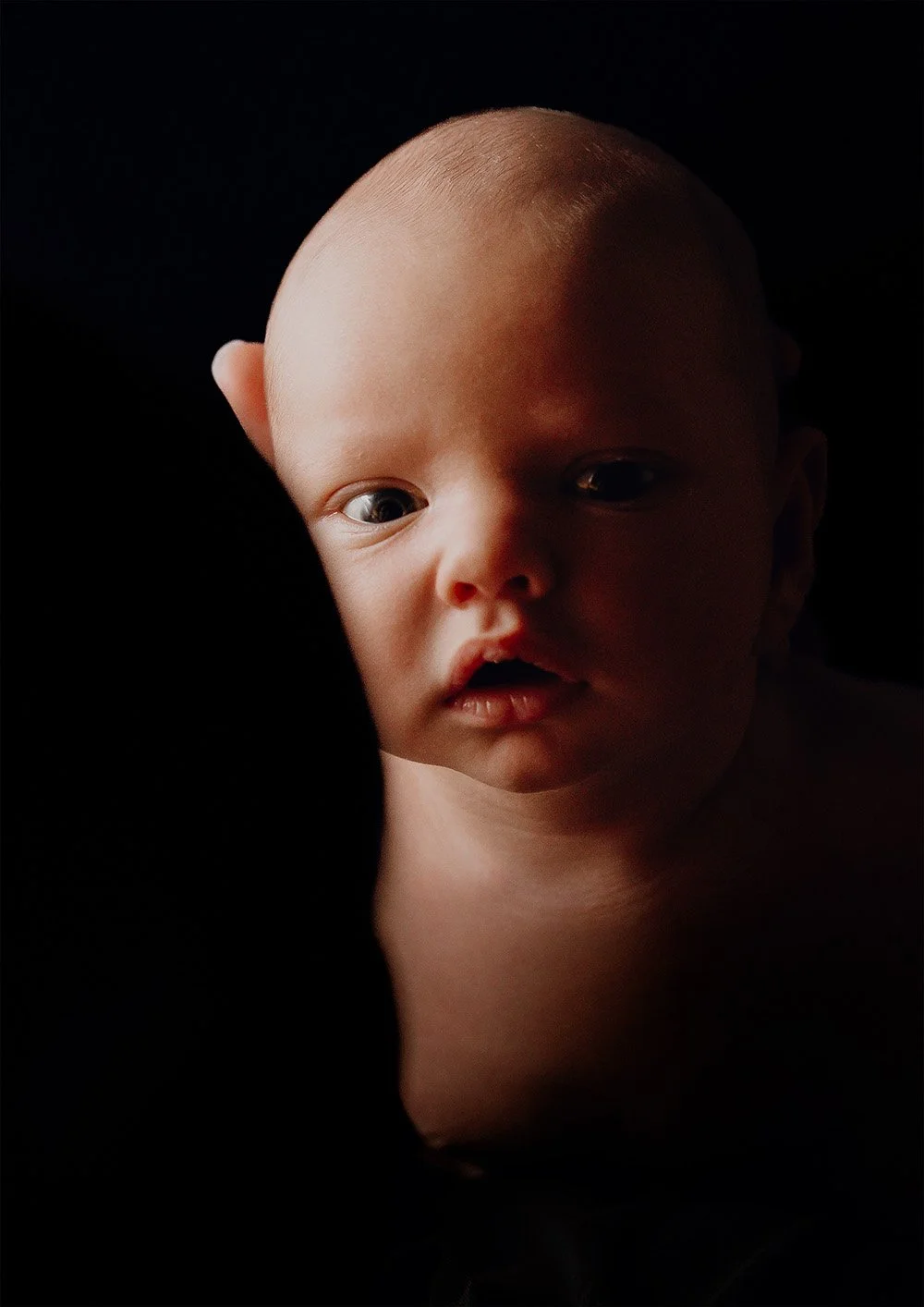 Nærbilde av en baby med lys hud og blå øyne, mot en mørk bakgrunn.