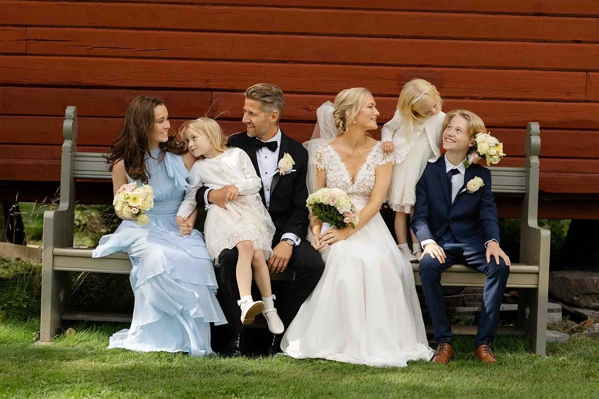 Et bryllupsbilde av en familie i formelle klær som sitter på en benk foran en trevegg, inkludert to voksne i brudekjole og dress og fire barn, alle smilende og holdende blomster.