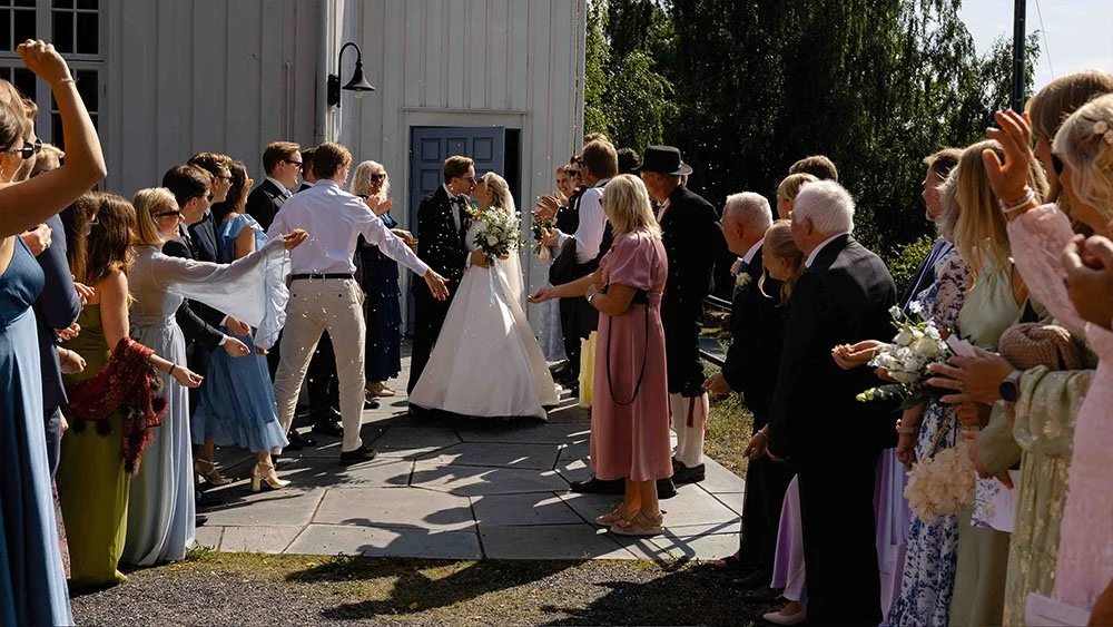 Et nygift brudepar går ut av Larkollen kirke, mens familie og venner kaster blomster utenfor kirketrappen på brudeparet. For flere slike bilder så se på porteføljen til vatafoto, som er en erfaren bryllupsfotograf i Moss.