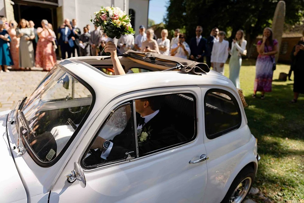 En brud med blomsterbukett stiger ut av en liten hvit bil i en hage, omgitt av gjester ved en utendørs bryllupsseremoni. Bildet er tatt i Gamlebyen i Fredrikstad.