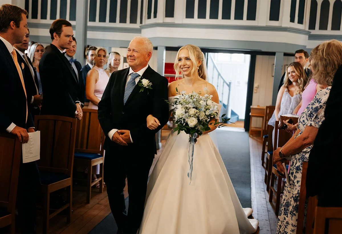 En brud går ned midtgangen i en kirke, ledsaget av en eldre mann, sannsynligvis faren, under en bryllupsseremoni. Bruden holder en stor blomsterbukett og smiler.