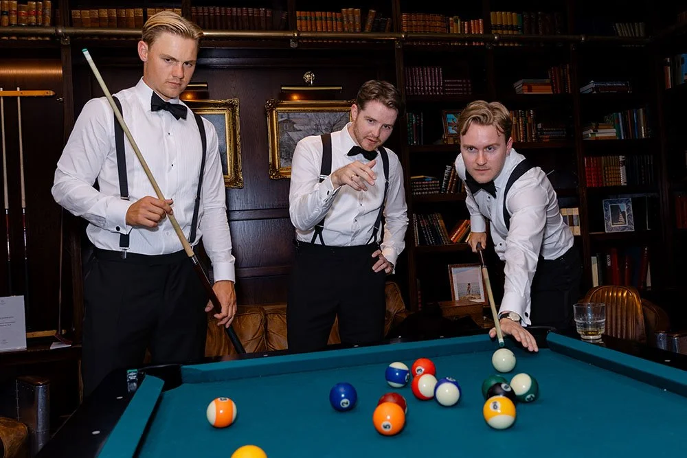 Tre menn i smoking spiller snooker i et rom med bøker og malerier på veggen.