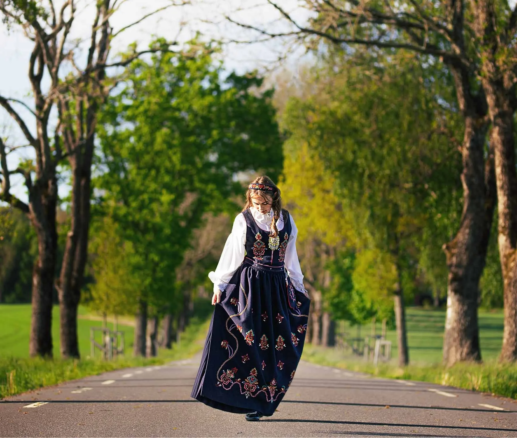 En kvinne i tradisjonell norsk bunad går langs en vei i en park med grønne trær. Det er vår eller sommer, og kvinnen har lang, blond hår og ser ned. Bakgrunnen er fylt med løvtunge trær og en blå himmel.