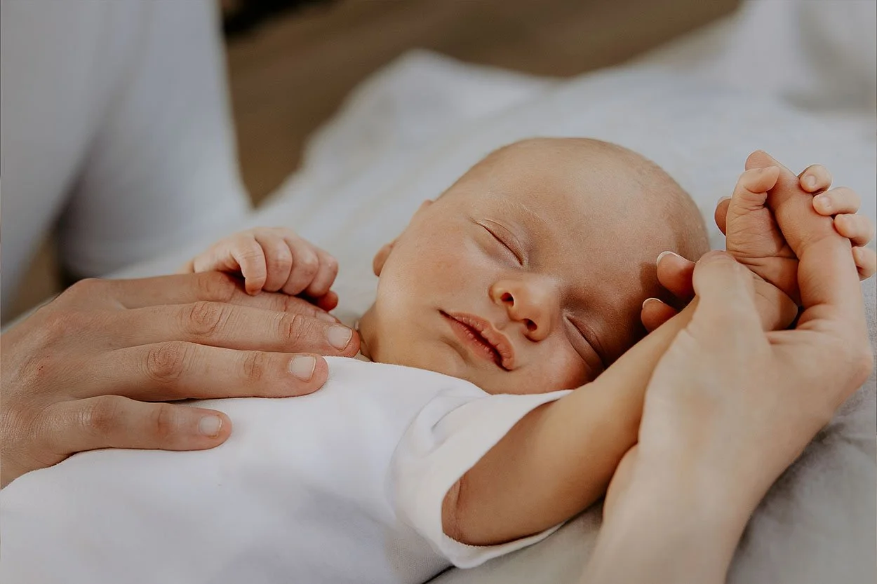 Et sovende baby med lukkede øyne holder en voksnes hånd. Bildet er tatt av fotograf Vatafoto, en erfaren fotograf som dekker alle Østfold-byene: Fredrikstad, Sarpsborg, Moss og Halden.