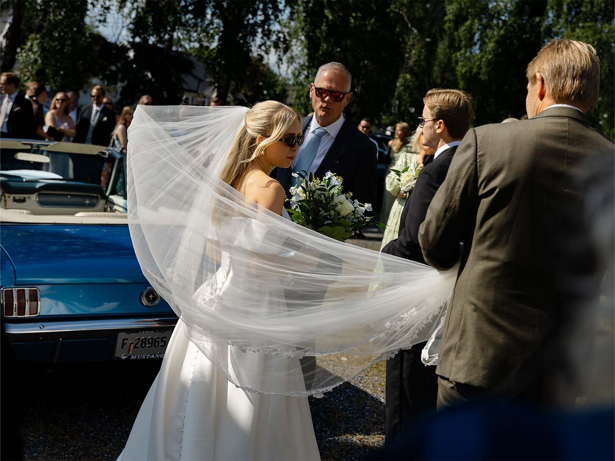 En kvinne i brudekjole og brudeslør, samt flere personer som ser ut til å delta i et bryllup, utenfor en klassisk bil