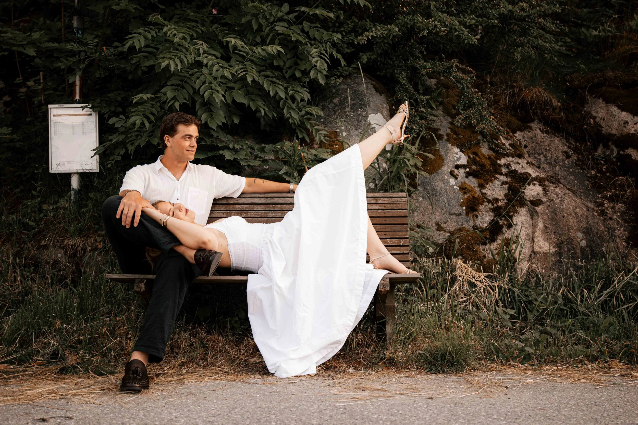 Et ungt par sitter på en trebenk i en park. Mannen, iført hvit skjorte og svarte bukser, holder en jente i en hvit kjole i armene, som ligger med øynene lukket. Jentas ene ben er hevet opp, og hun har på sandaler. Bakgrunnen er fylt med grønt løv og 