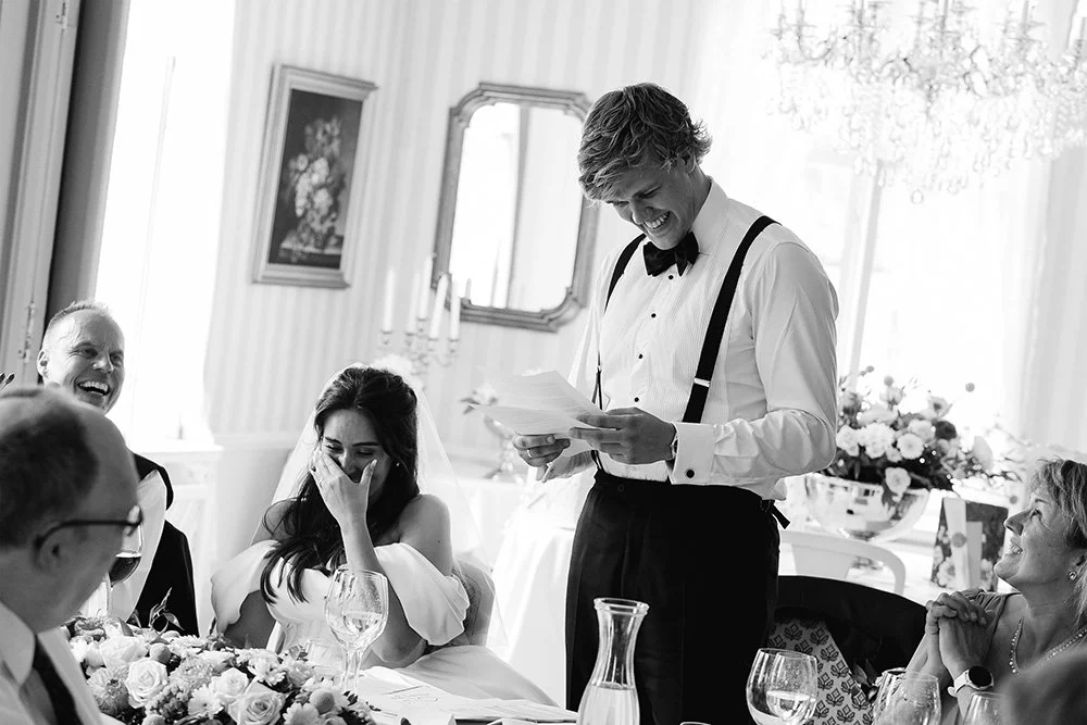 En man holder en tale under et bryllup, omgitt av glade gjester som ler og ler, i et elegant rom med blomster og dekorasjoner.
