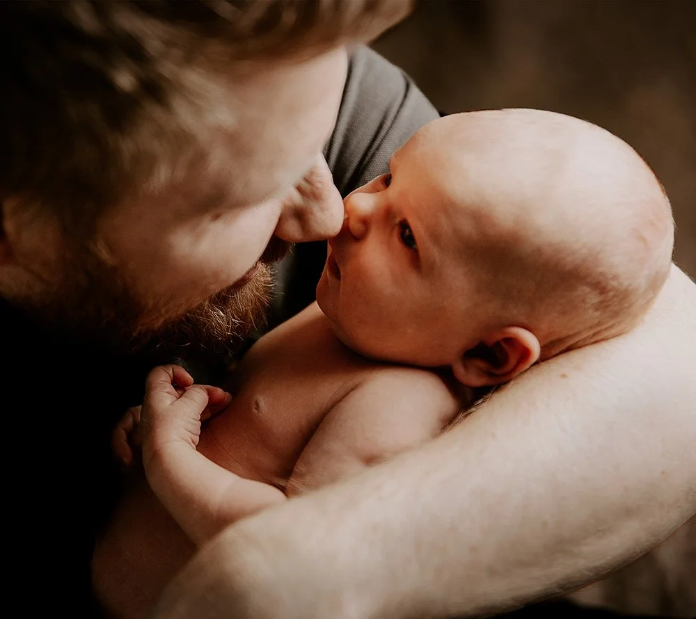 En voksen mann med skjegg holder en spedbarn, som ligger på hans armer, tett inntil ansiktet hans, begge ser på hverandre. Bildet er eksempel på hva man få når man bestiller nyfødtfotografering av Vatafoto.