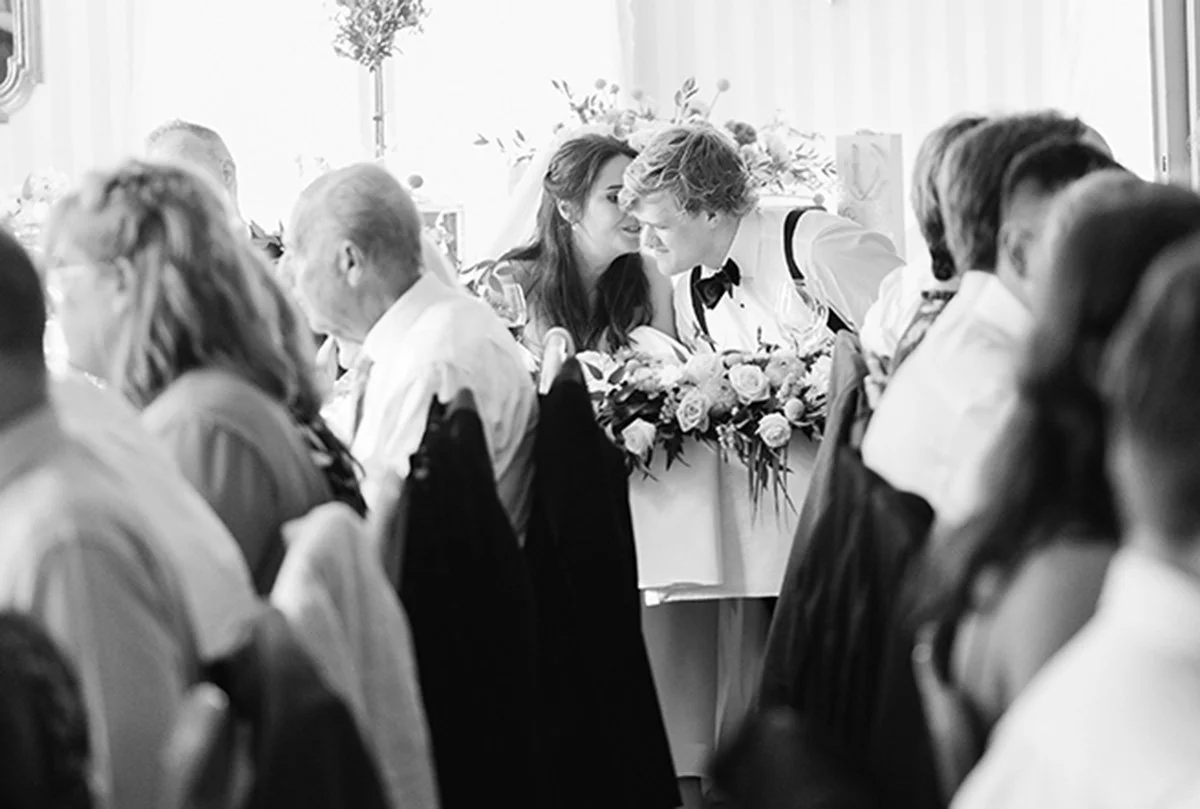 To mennesker hvisker nære hverandre ved et bord med blomster, omgitt av flere personer, i en svart-hvitt bilde.