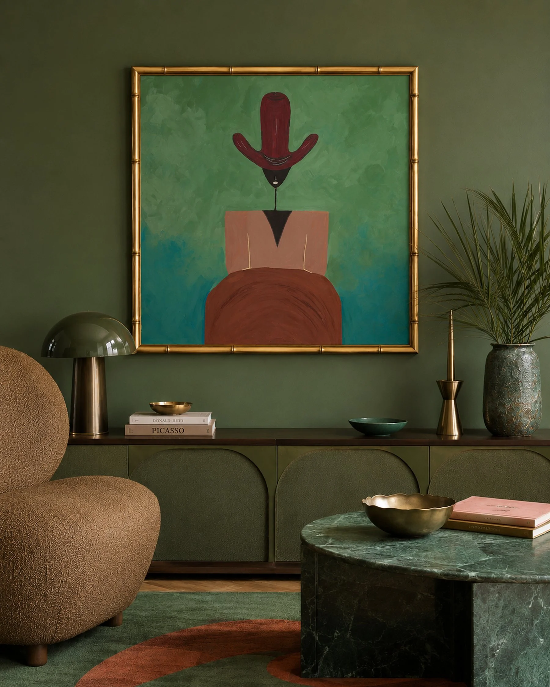 A living room with green walls decorated with a painting of minimalistic abstract art featuring a red hat and black geometric shapes. The living room has a brown upholstered chair, green carpet, gold-framed lamp, green marble coffee table, a large va