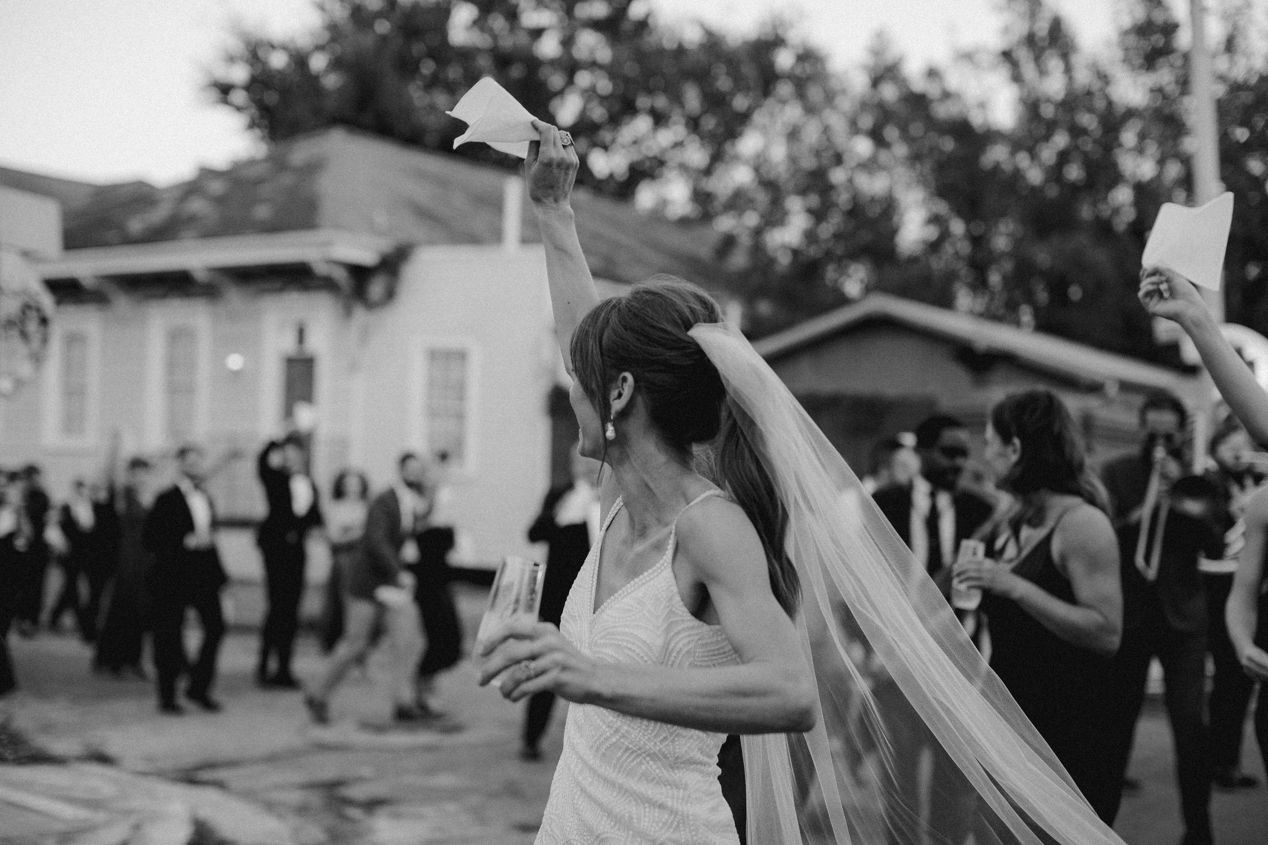 Black and white photo of a bride with a veil dancing at her wedding reception, holding a tissue in one hand while other guests dance in the background.