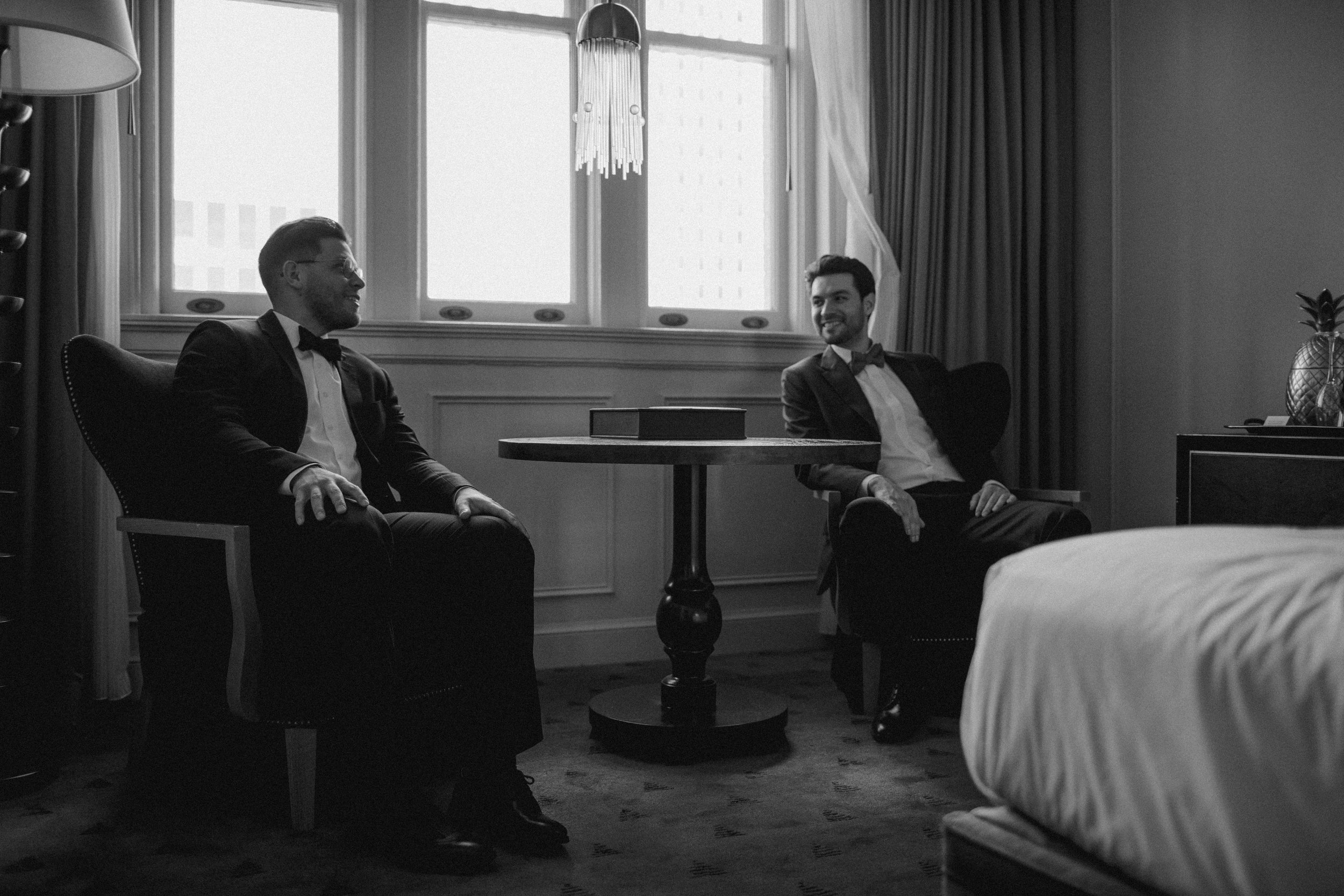 Two men in tuxedos sit and chat in a hotel room, with a window and curtains in the background, and a bed partially visible in the foreground.