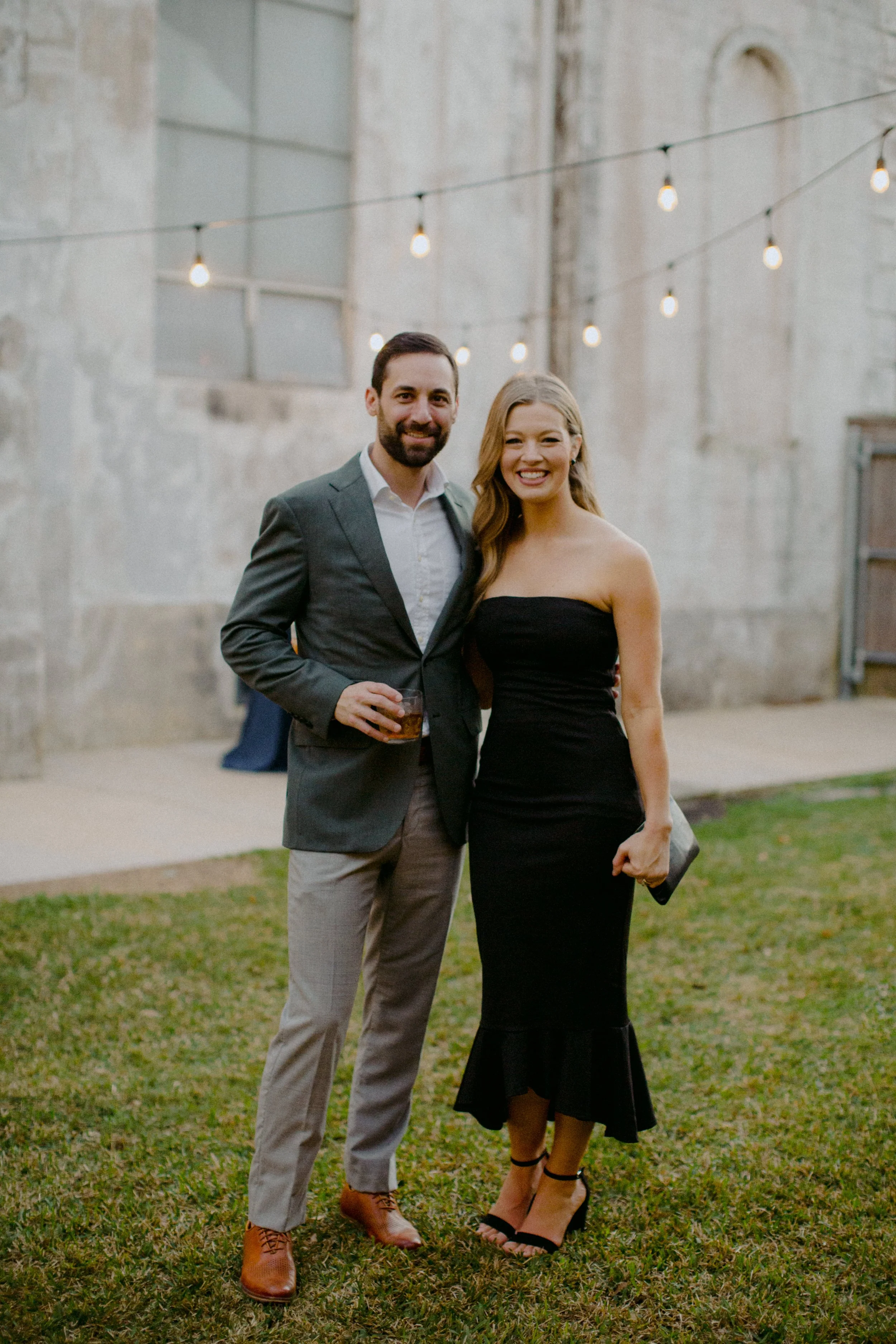 A man and woman smiling and standing outdoors in a yard with string lights hanging above them, dressed in formal attire.