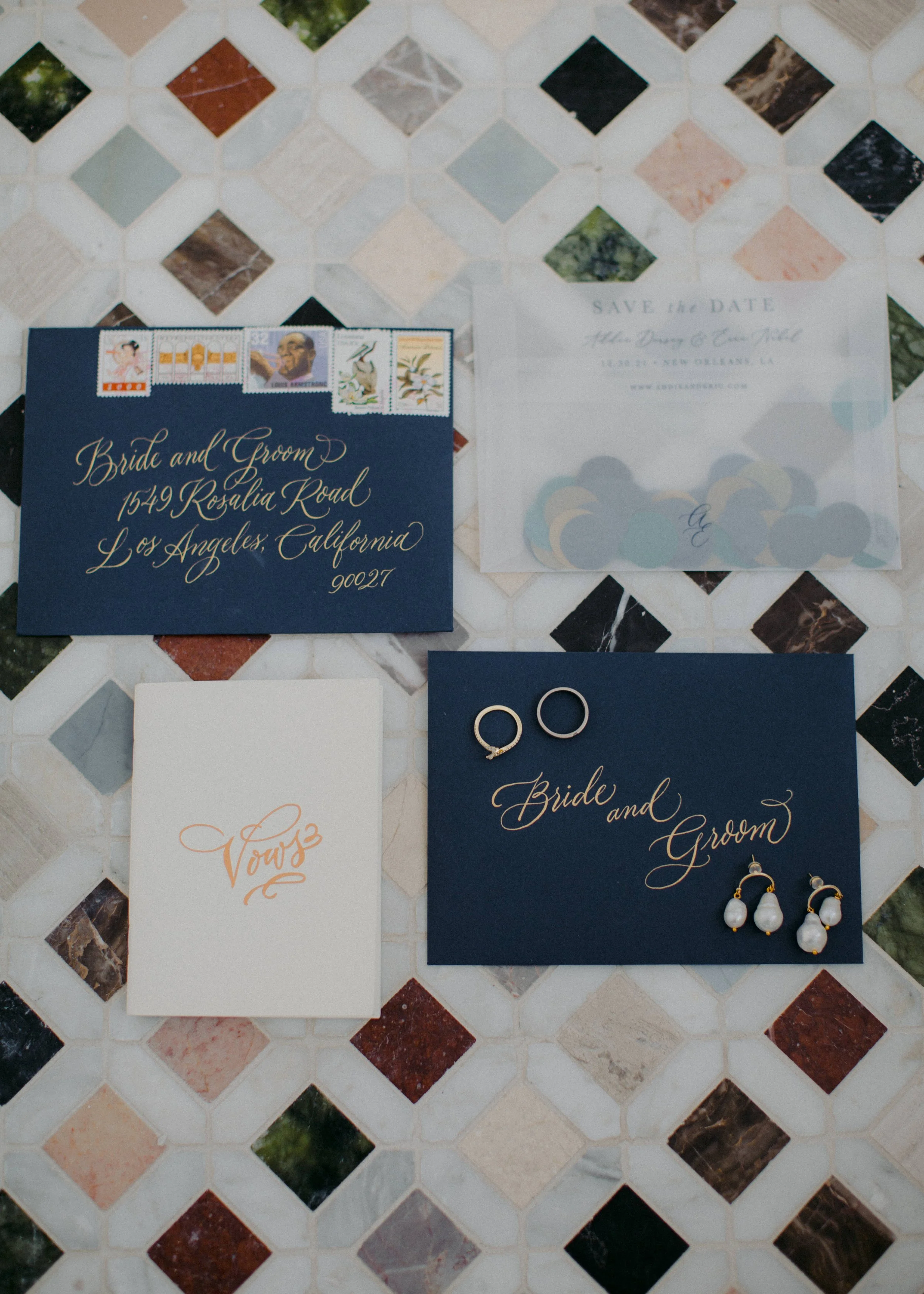 Wedding invitation suite including a navy envelope addressed to the bride and groom, postage stamps, a clear 'Save the Date' card, a white vow card, and a navy card with gold lettering holding wedding rings and earrings, all displayed on a tiled surf