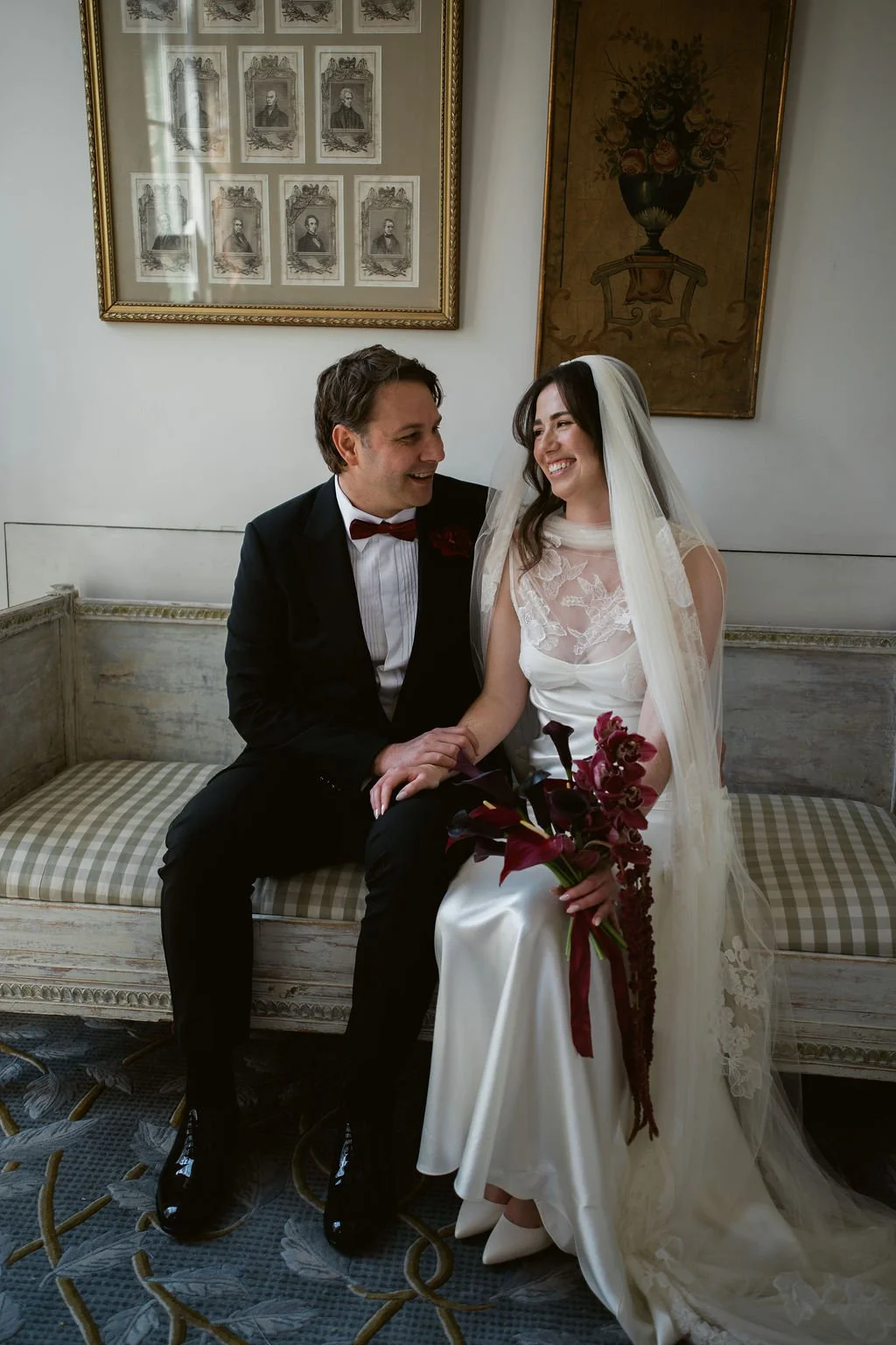 Couple sitting on a sofa woman is wearing a white dress and veil and holding a burgundy bouquet