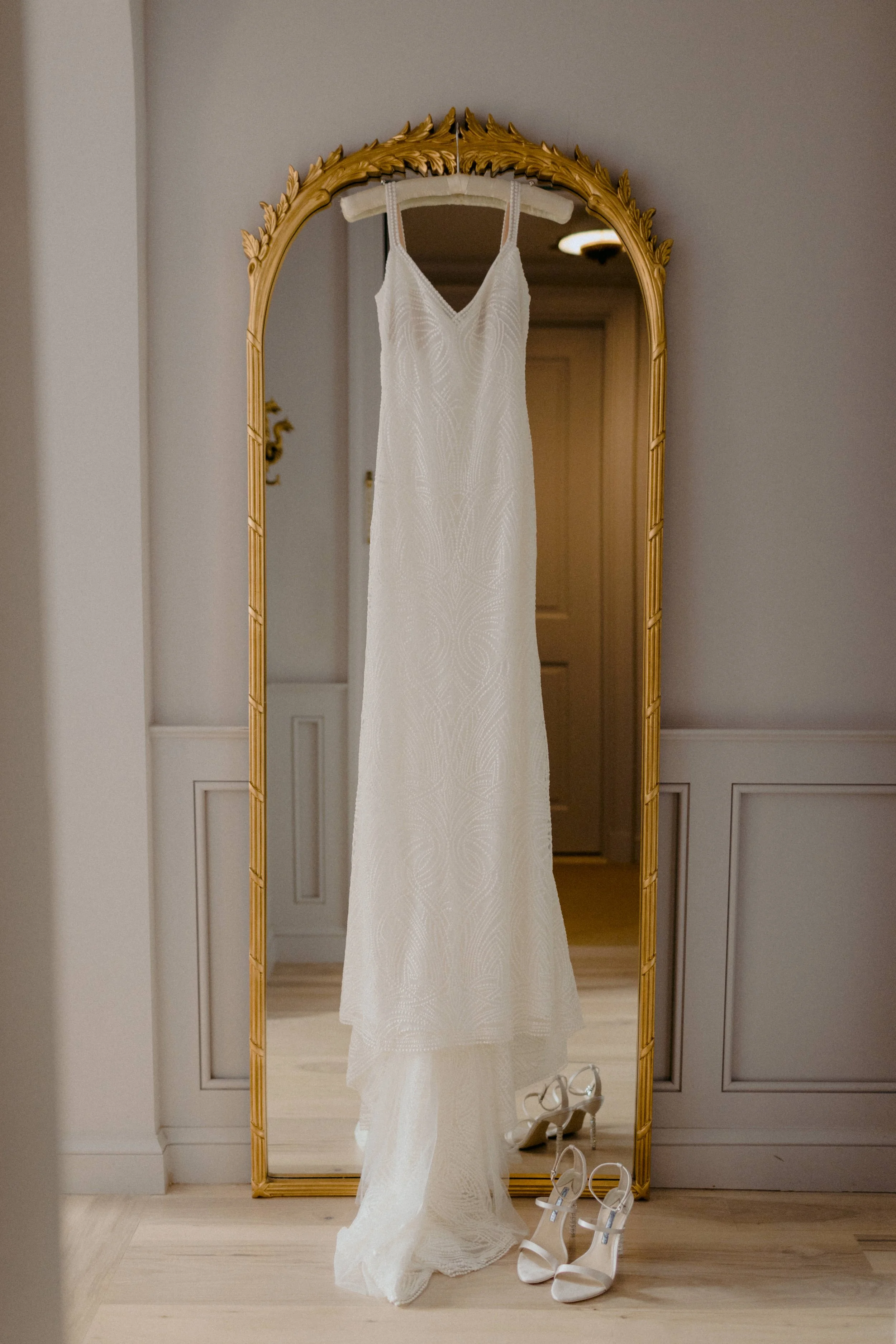 A white wedding dress hanging on a wall-mounted hanger in front of a tall, ornate gold-framed mirror. A pair of white high-heeled shoes are placed on the floor below the dress.