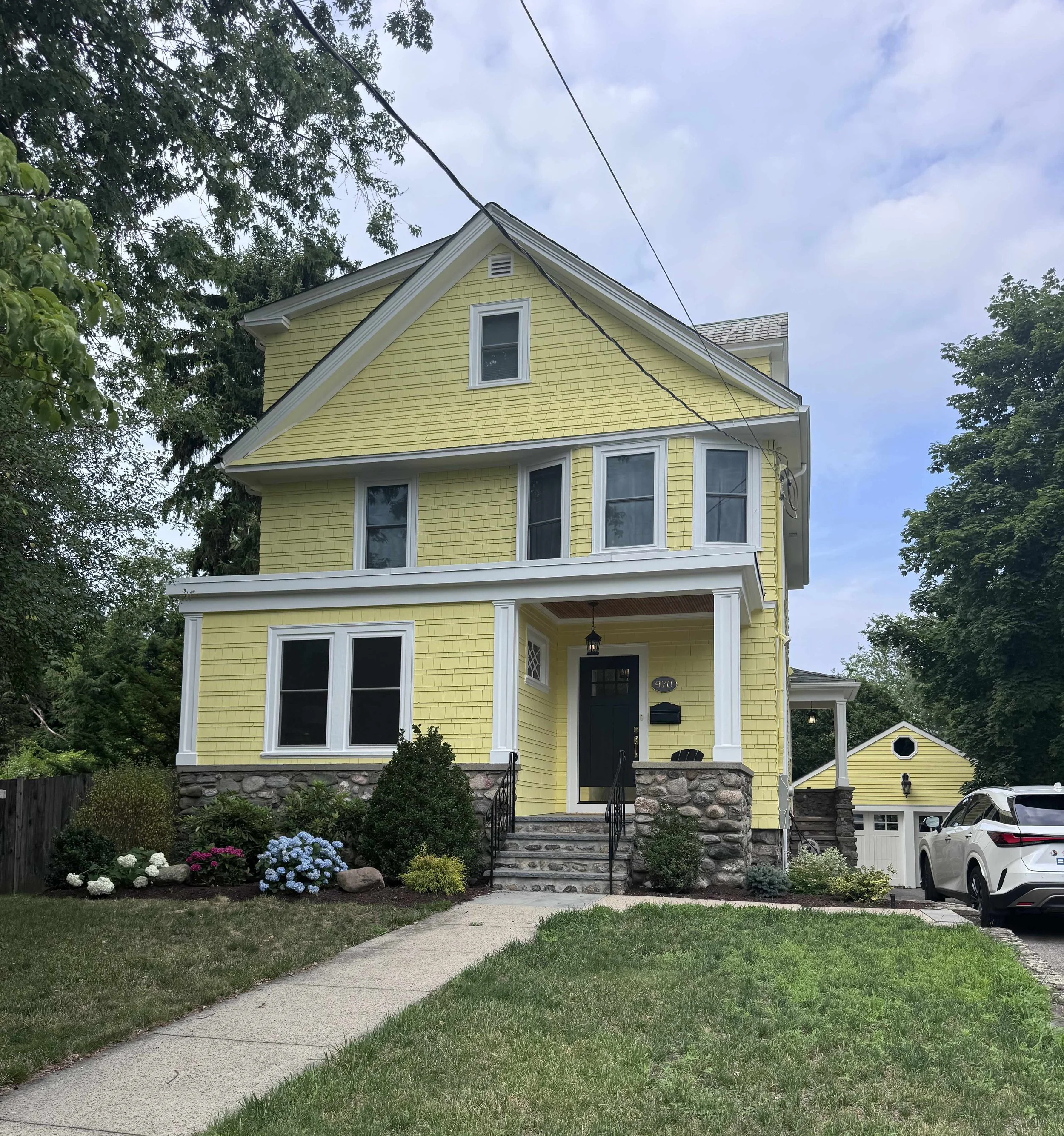 Yellow three-story house with white trim, stone steps, a small front porch, and a black front door, surrounded by a well-maintained lawn, flower beds with hydrangeas, trees, and a sidewalk in a suburban neighborhood during the day with partly cloudy sky.