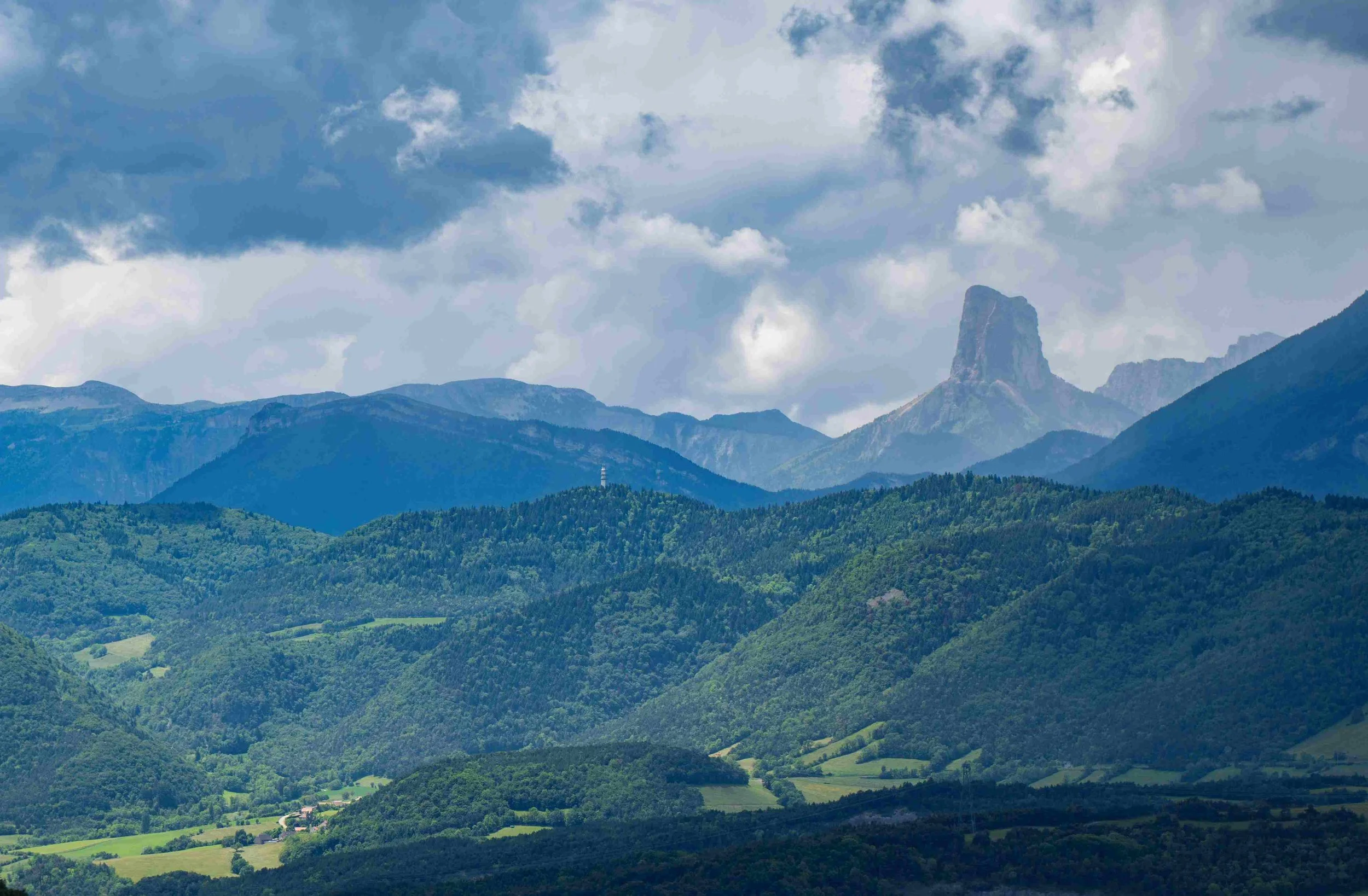 Scenic view of green forested mountains with a distinct rock formation peak in the background under cloudy sky.