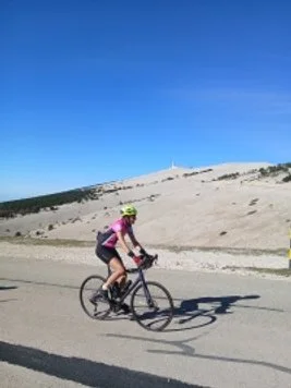 Cyclist riding a bike on a mountain road with a sandy hillside and clear blue sky in the background.