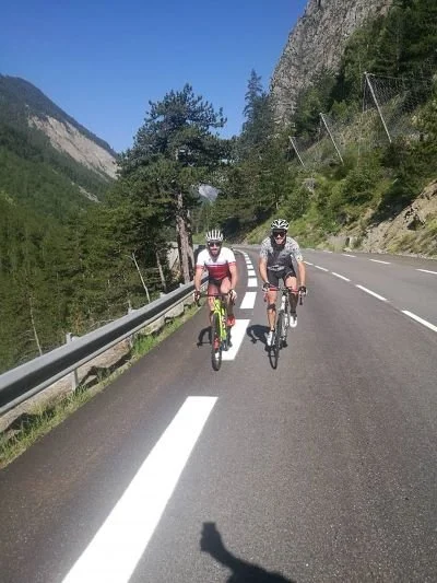 Two cyclists riding on a mountain road with a guardrail, trees, and rocky cliffs in the background, under a clear blue sky.