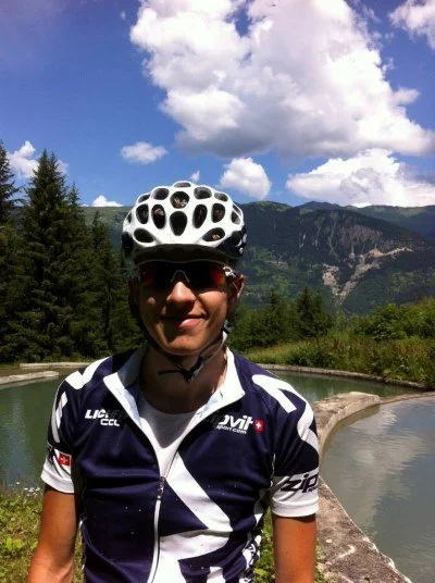 A man wearing a white cycling helmet, sunglasses, and a navy blue cycling jersey with white accents, standing outdoors near a water feature with mountains, trees, and a partly cloudy sky in the background.