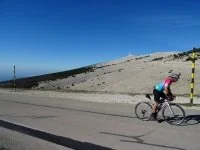 A cyclist wearing a helmet and colorful shirt rides a purple bicycle on a paved road with a mountain landscape in the background, featuring a summit with a communication tower.