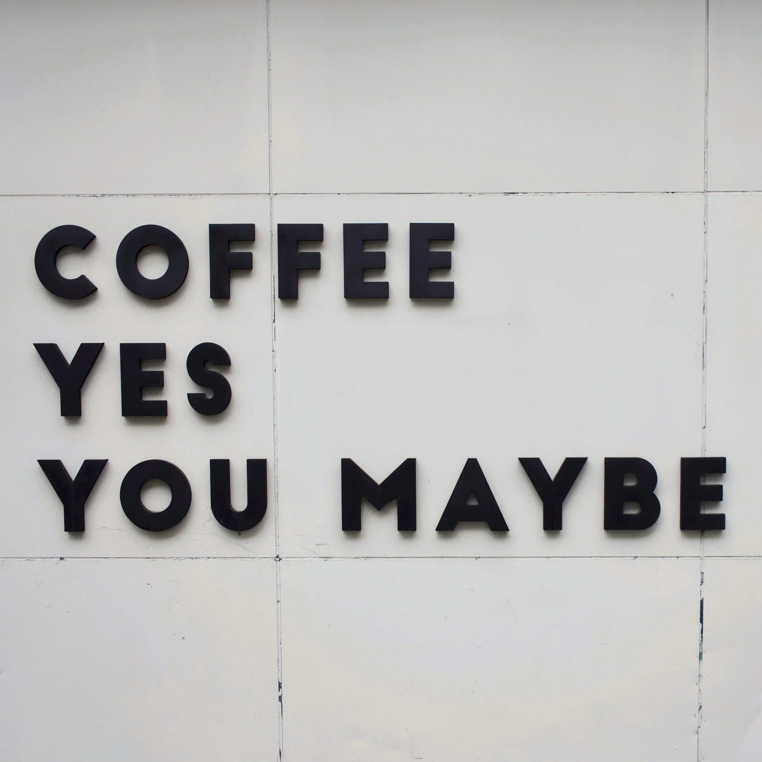 Sign on a white wall with black 3D lettering that reads: 'COFFEE YES YOU MAYBE'.