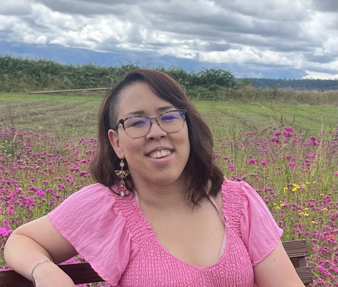A therapist in a pink summer dress with ruffled sleeves, sitting in nature on a bench in a field of pink and purple wildflowers, under a cloudy sky.