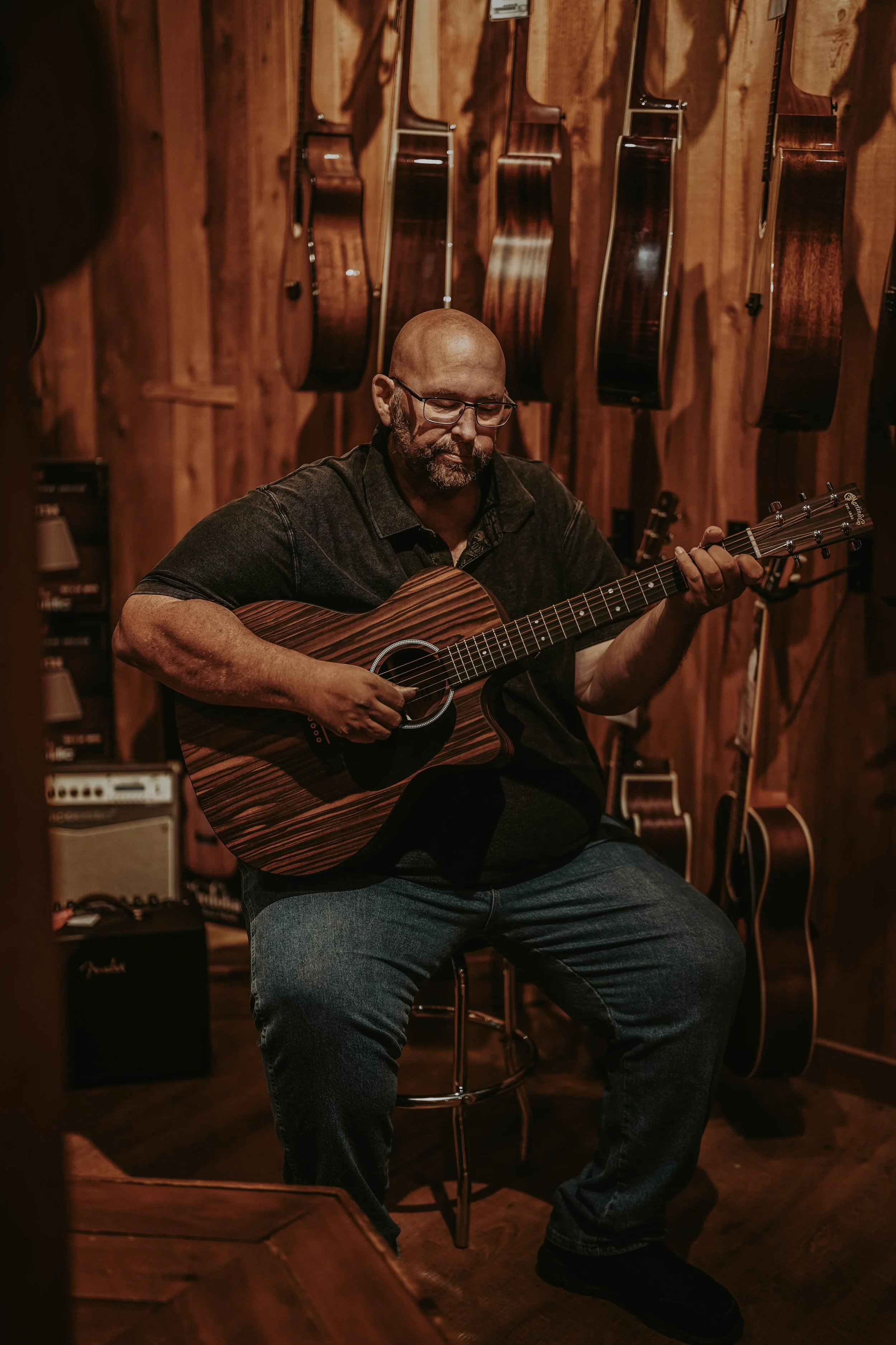 A man with glasses and a beard playing an acoustic guitar in a wood-paneled music store, surrounded by hanging guitars and musical equipment.