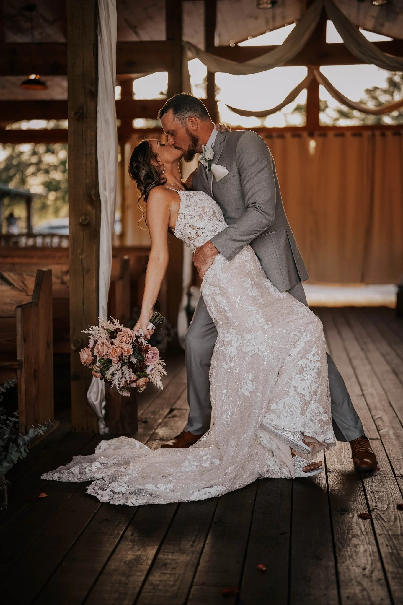A bride and groom share a kiss during their wedding ceremony inside a rustic wooden venue. The bride wears a lace wedding gown and holds a bouquet of pink roses, while the groom is dressed in a light gray suit with a boutonniere. The setting has natural light and wooden decor.