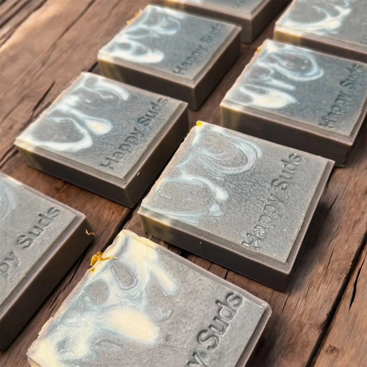 Several handmade soap bars with a marbled gray, white, and beige pattern, embossed with the words "SPRS" and "HAPPY DAY SPRS", arranged on a wooden surface.
