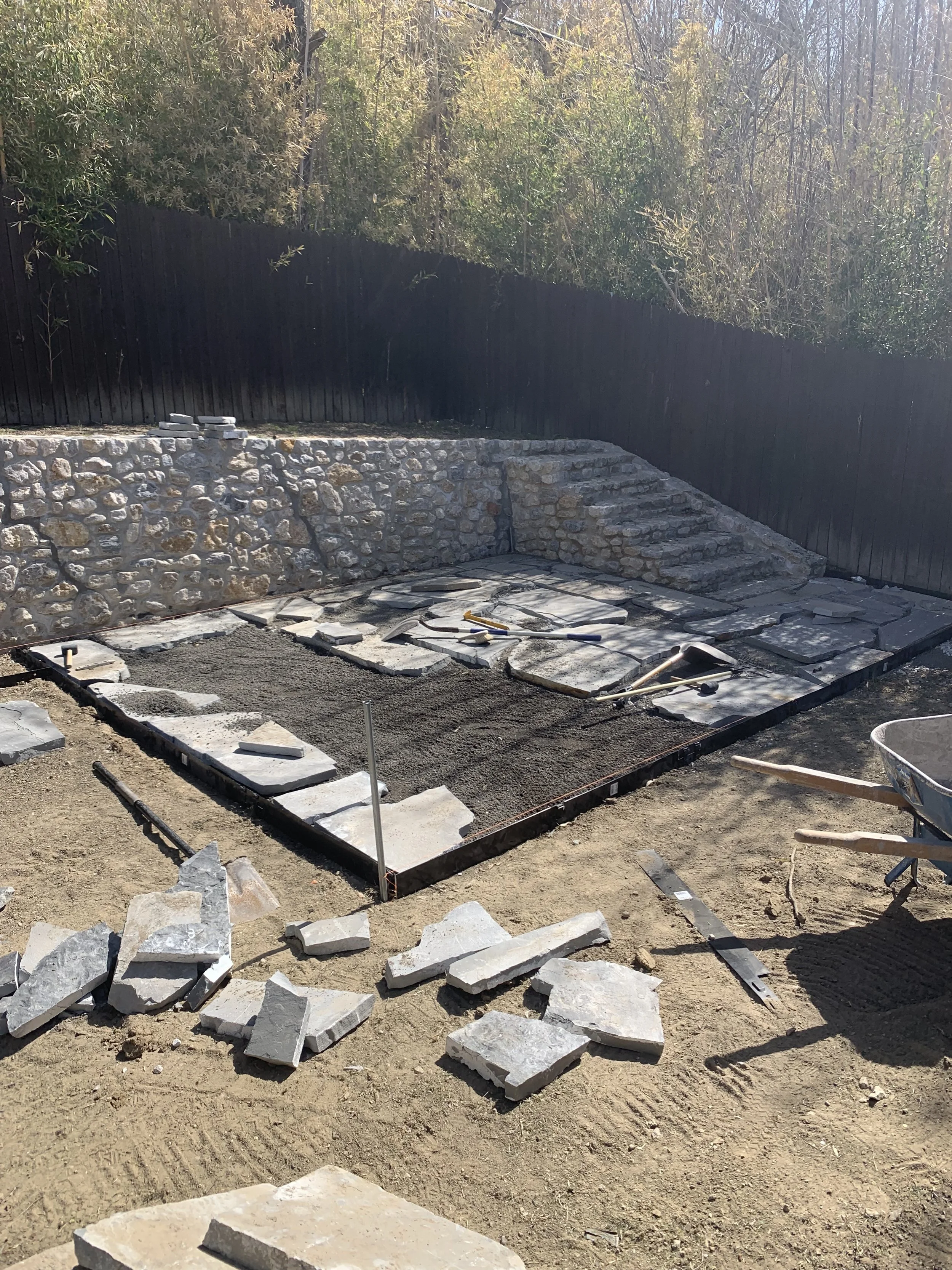 Construction site with stone pavers and gravel being laid for a patio or walkway, with tools and materials around.