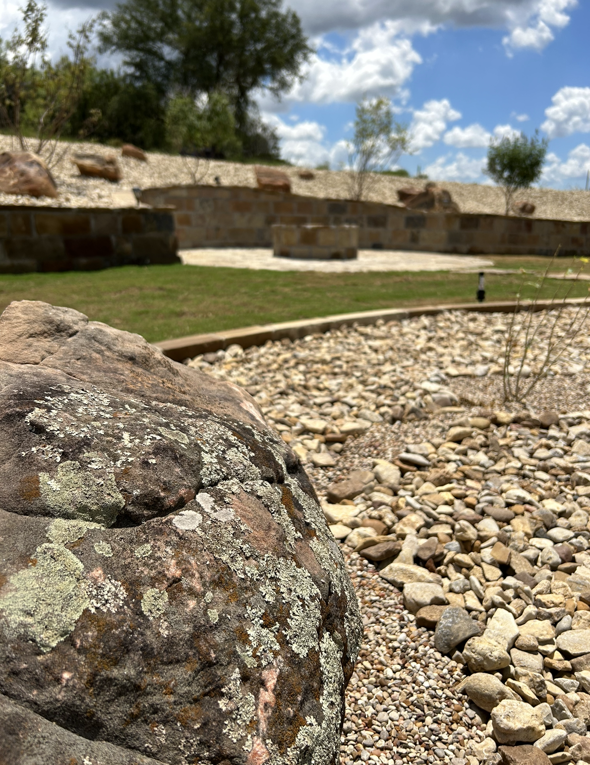Close-up of a large rock in a landscaped yard with gravel, grass, trees, and a brick wall in the background under a partly cloudy sky.
