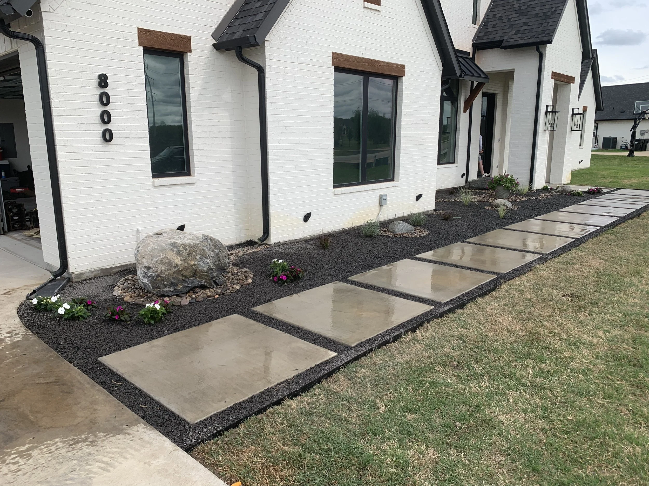 Concrete stepping stones leading to the front door of a white brick house. The house has black trim and installed black gutters. There are small landscaped flower beds with rocks, flowers, and plants along the pathway. A person is seen near the entrance, and there is a grassy lawn extending beyond the pathway.